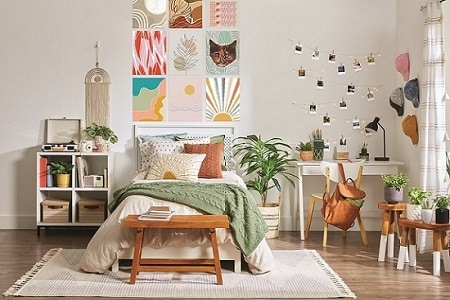 Cozy bedroom with a neatly made bed, pillows, and a green throw. Art prints hang above the bed with Command Strips. A desk, chair, and photos with Command Hooks add charm. Plants enhance warmth.
