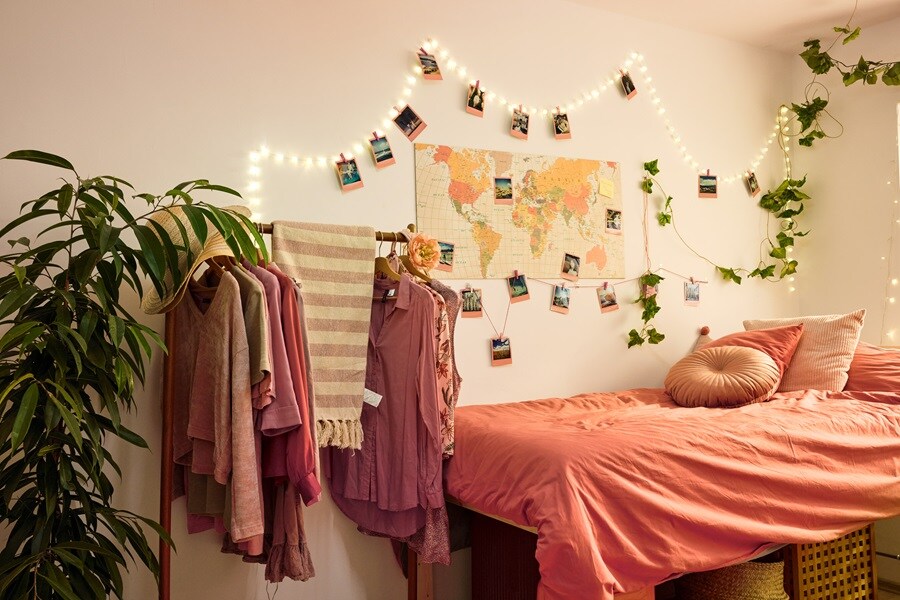 Student bedroom wall with fairy lights, photos, a world map and plant branches hung using Command Clear Hooks, plus a tidy bed and clothes rail.
