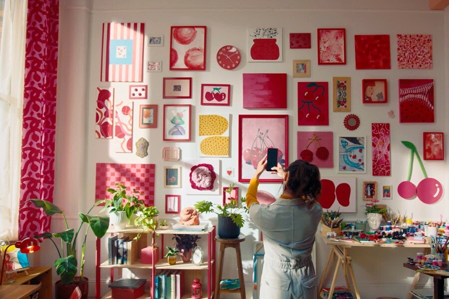 Living room with cherry‑themed art hung using Command Strips, plus a shelf of books and plants and a table with art supplies.

