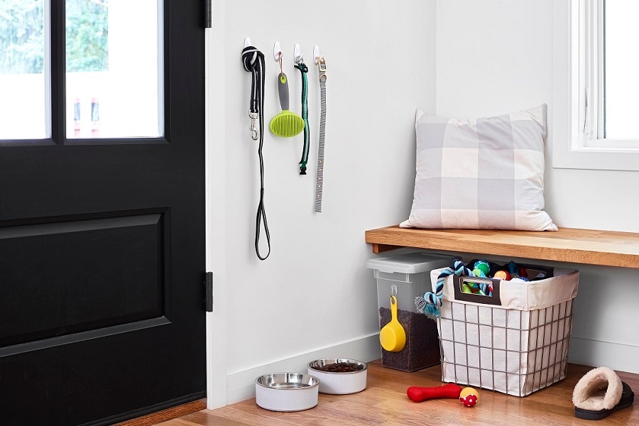 Tidy entryway with Command hooks holding a dog leash, brush, and collar. Below are pet bowls, a storage bin, and a toy basket. A cushioned bench sits above the storage area.
